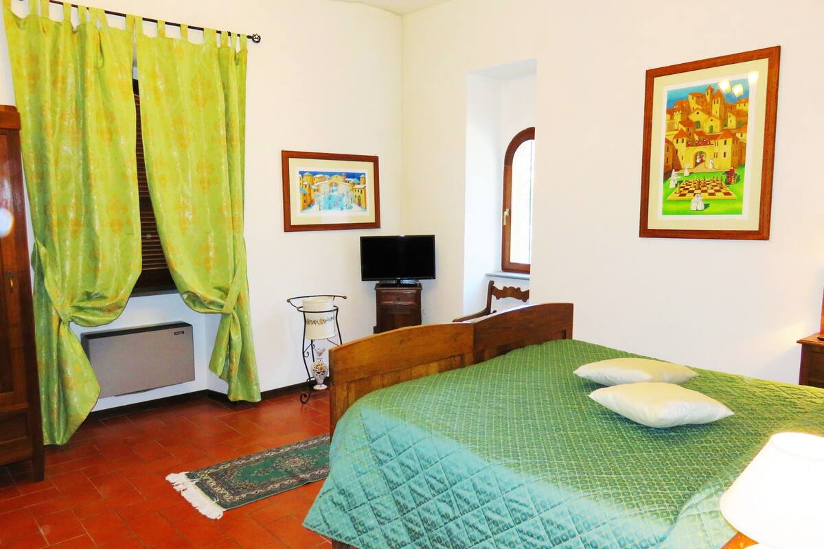 Un dormitorio con cama, televisión, ventana con cortinas, un cuadro enmarcado en la pared y suelo de baldosas.