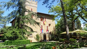 A historic stone building with a tower, surrounded by lush greenery and outdoor seating. - Castello Cortevecchio (Gubbio)