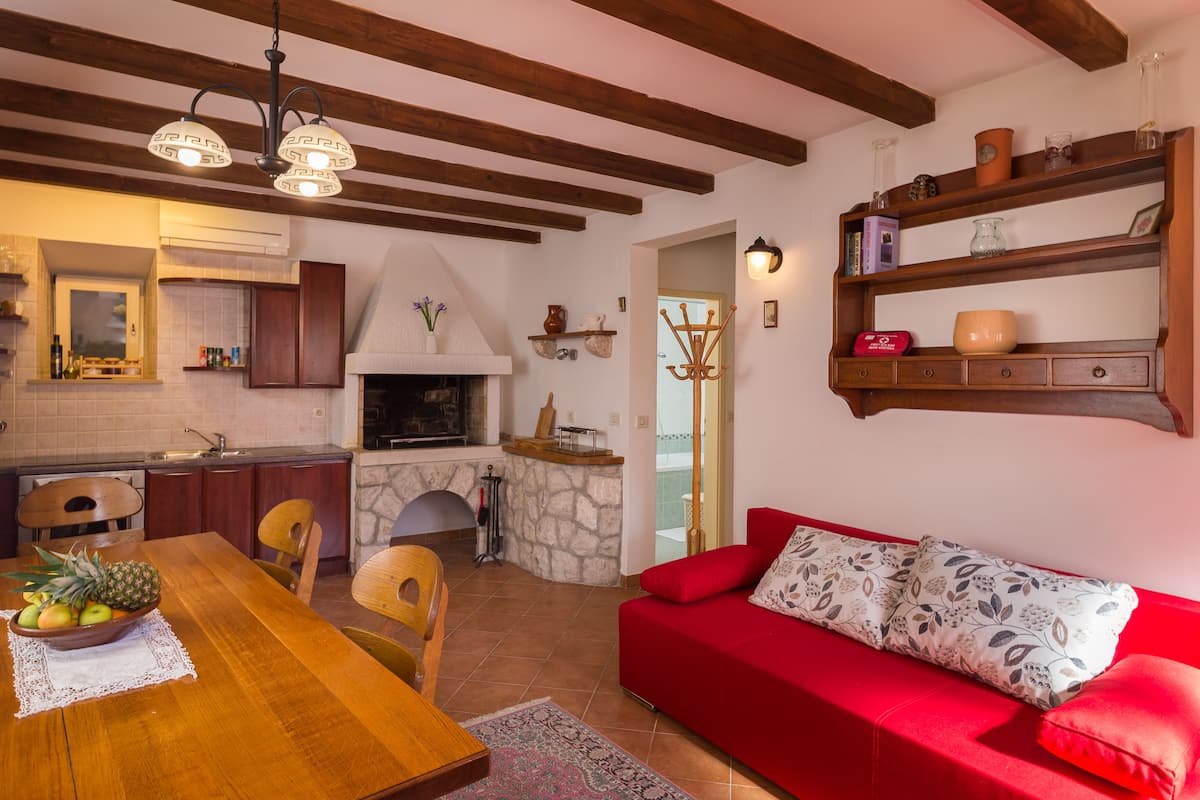 A cozy living room with a red sofa, wooden dining table with fruit, and a stone fireplace.