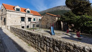 A stone building with a red-tiled roof, a playground with a slide, and a stone wall. - Apartments Vi-La (Dubrovnik)