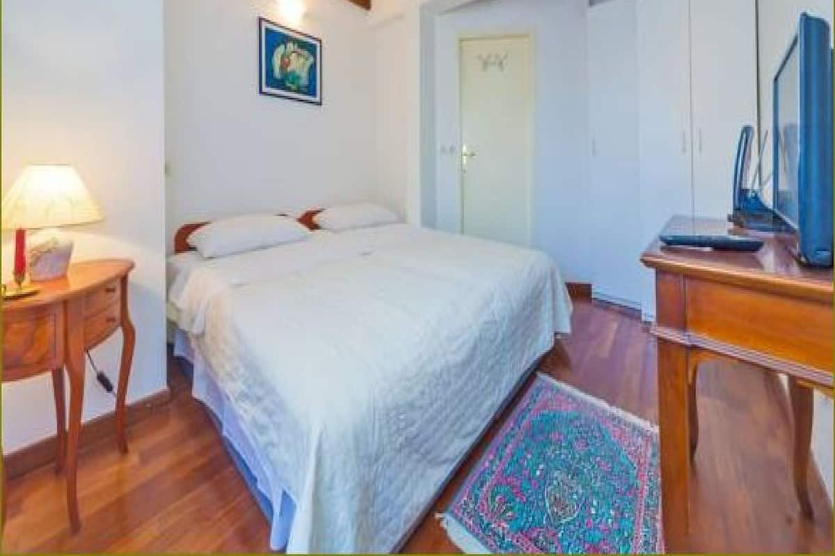 A bedroom with a bed, a nightstand, a desk, a chair, a door, and a wall-mounted TV.