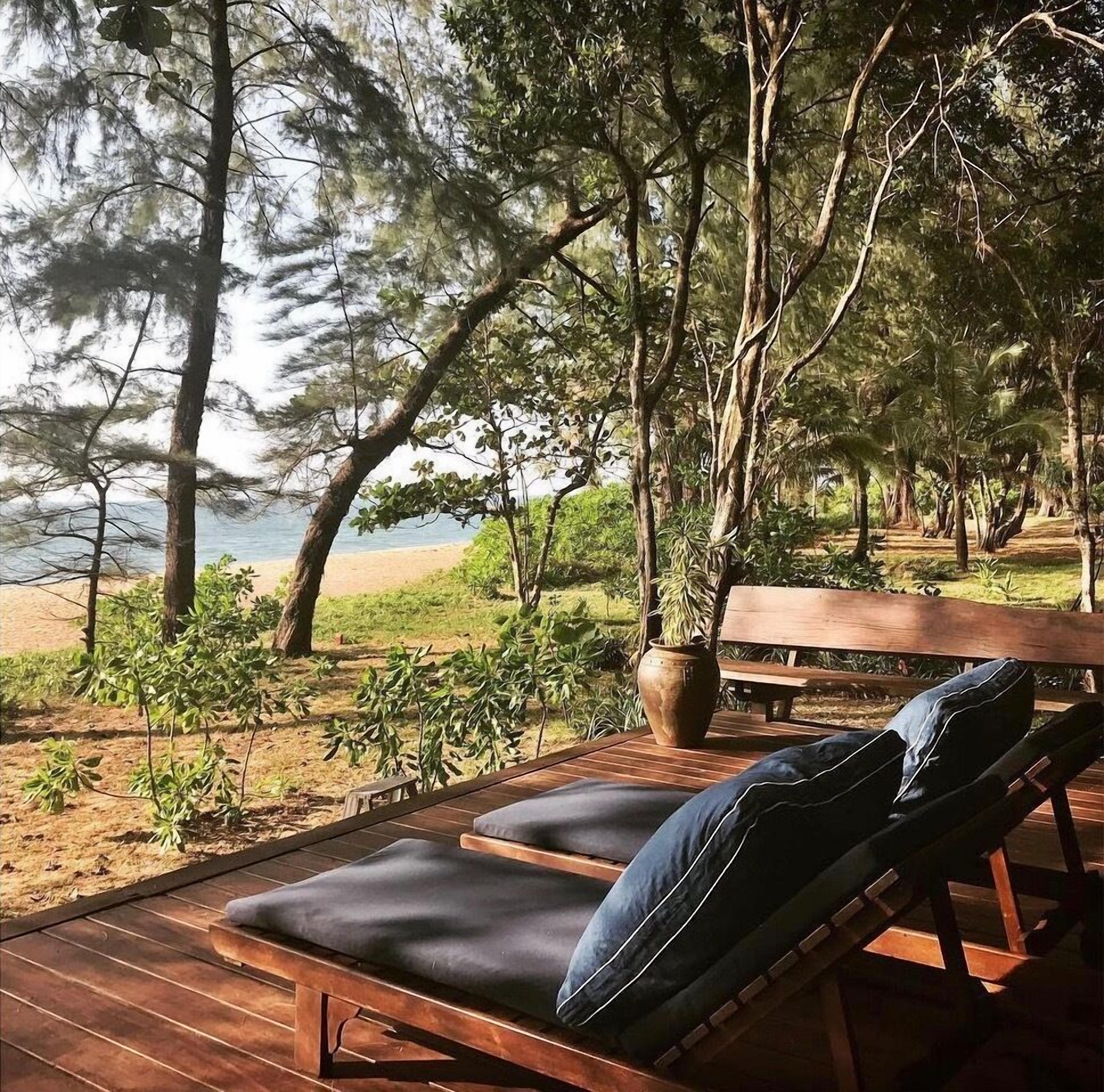 Una terraza de madera con dos sillones de relax y un banco, con vista a una playa con árboles y al mar.