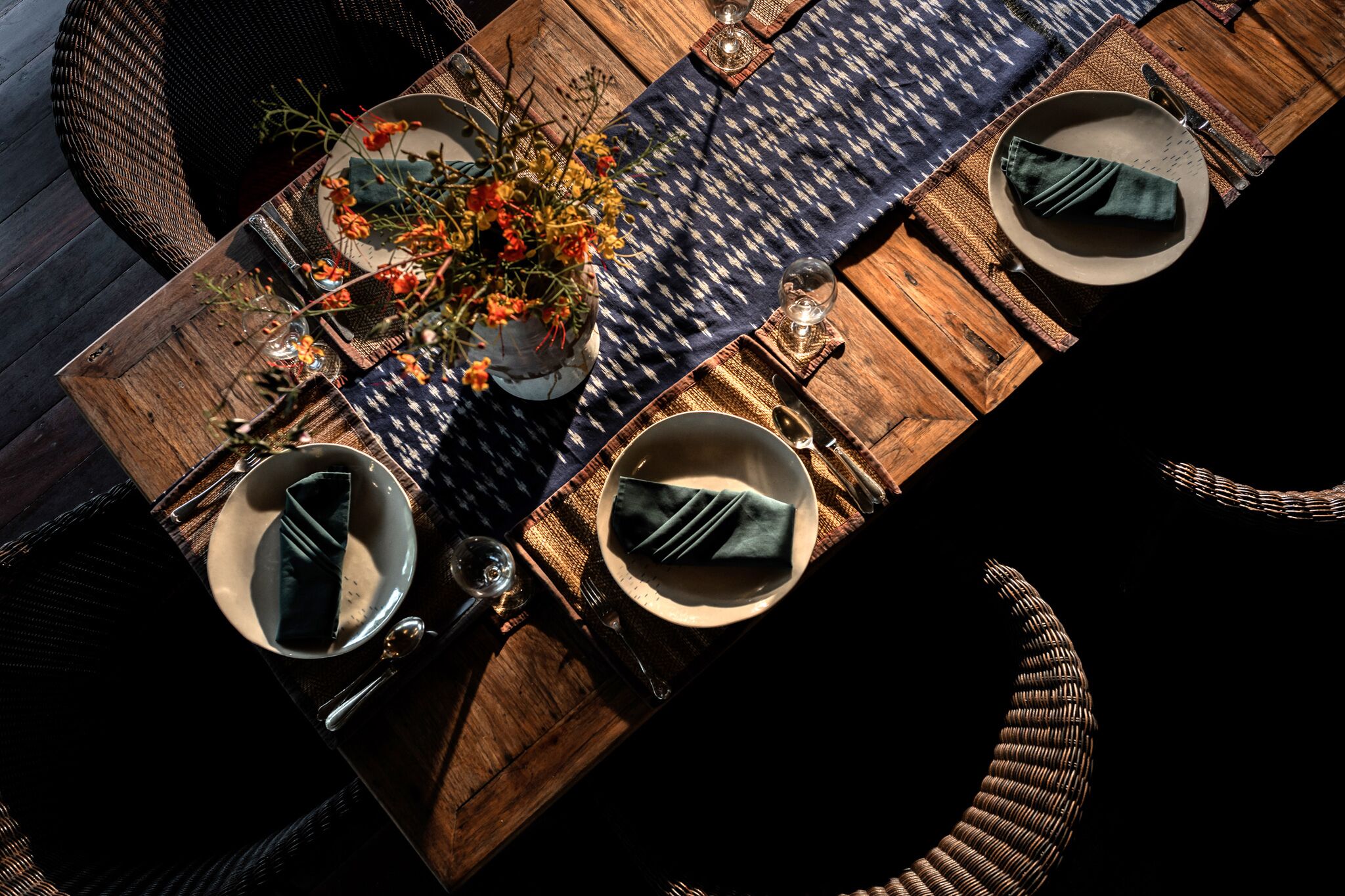 Een houten eettafel met een geweven placemat, twee borden met gevouwen servetten, een vaas met bloemen en een glas water.
