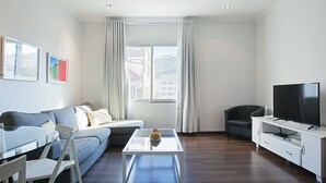 Un salon moderne avec un canapé, une table basse en verre, un téléviseur et une vue sur les immeubles par la fenêtre.