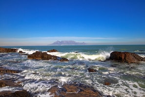 A rocky coastline with waves crashing against the rocks and a distant mountain range under a clear blue sky.