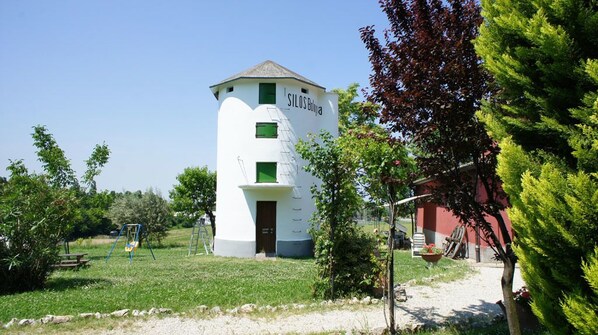 Un edificio cilíndrico blanco con una puerta y ventanas, rodeado de vegetación y un edificio rojo al fondo.
