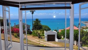 A view from a room with large windows overlooking a coastal area, including a road, greenery, and a small building. - Admirals Rest Self-Catering House (Mossel Bay)