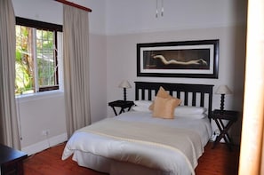 A neatly arranged bedroom with a large bed, two bedside tables with lamps, a framed artwork above the bed, and a window with curtains. - Admirals Rest Self-Catering House (Mossel Bay)