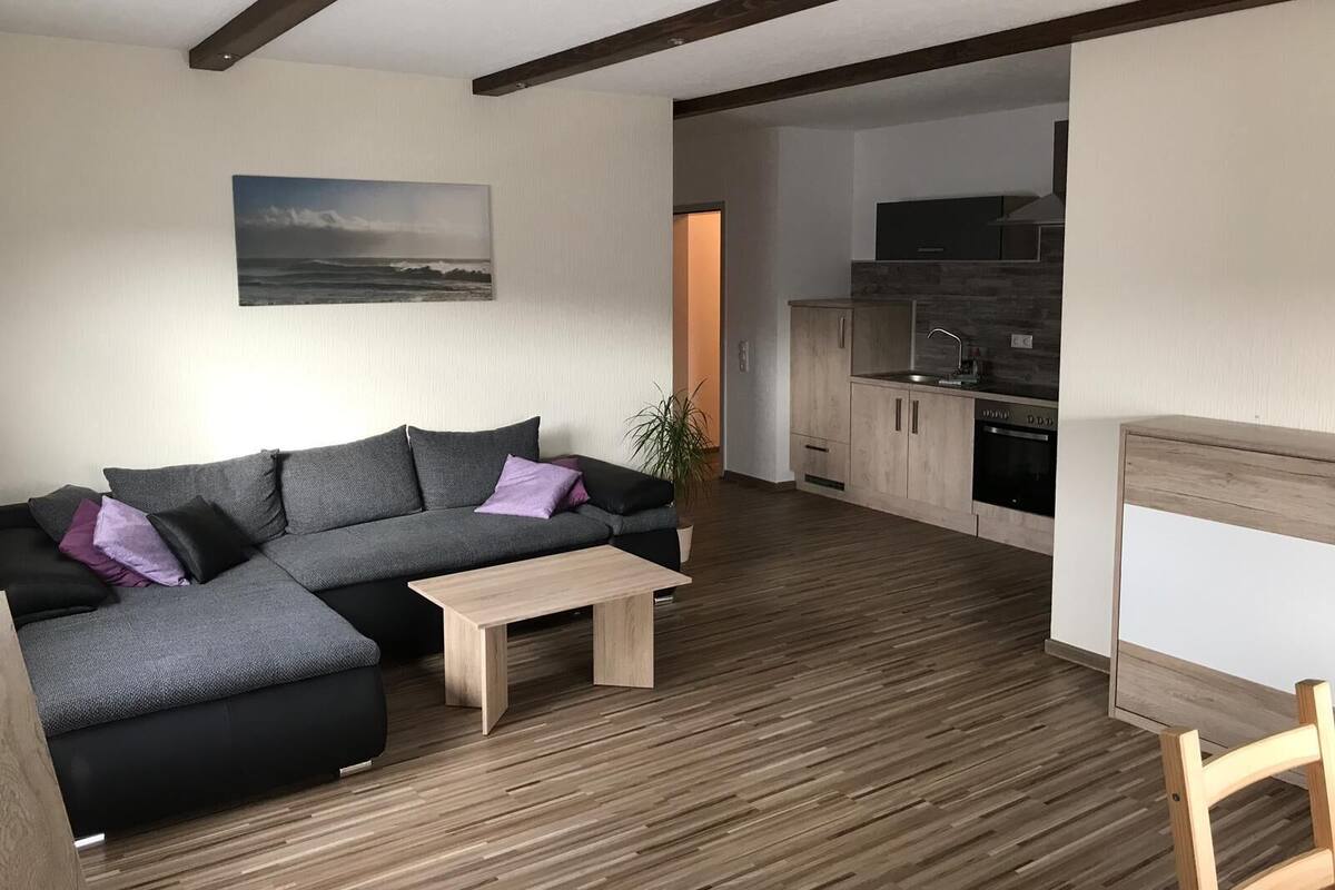 Una sala moderna con un sofá seccional gris, una mesita de centro de madera y un cuadro de un paisaje en la pared.
