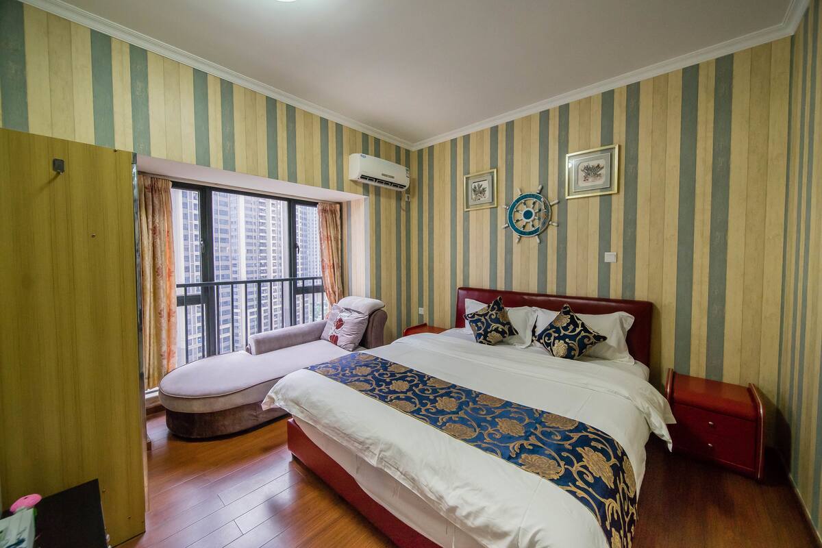 A hotel room with a large bed, a sofa, a window with curtains, and a wall-mounted air conditioner.