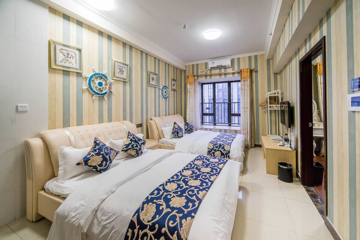 A hotel room with two beds, striped wallpaper, a desk, and a window with curtains.