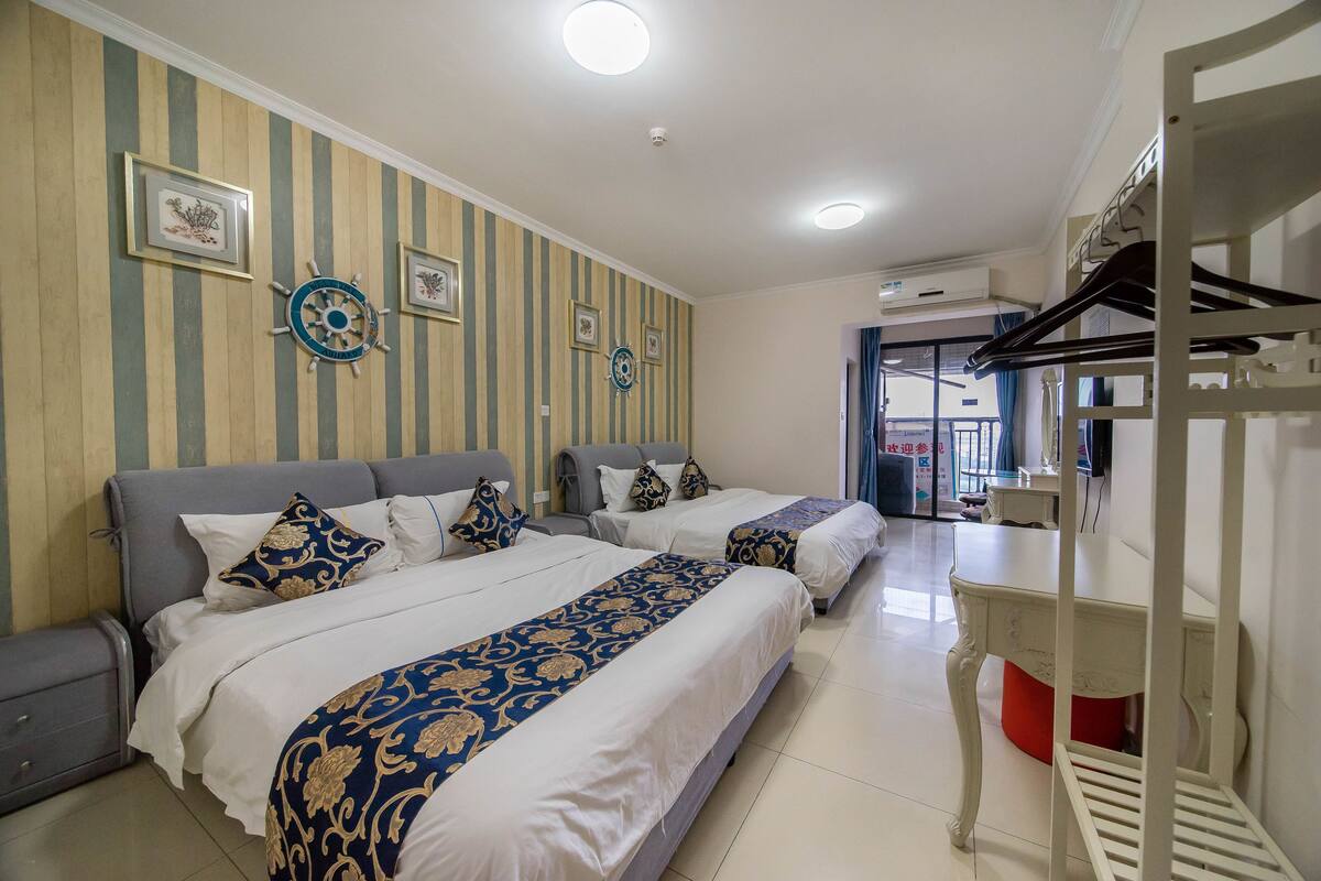 A hotel room with three beds, striped wallpaper, decorative wall art, a small table, and a red stool.