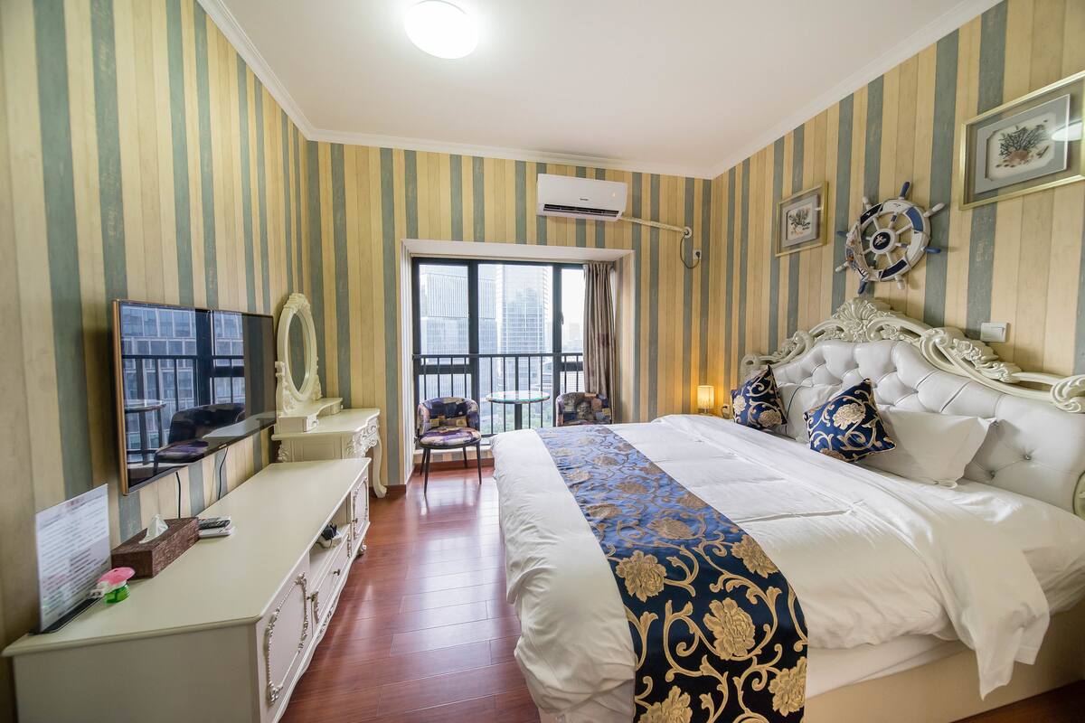 A hotel room with a large bed, a TV, a desk, and a balcony with a view of buildings.