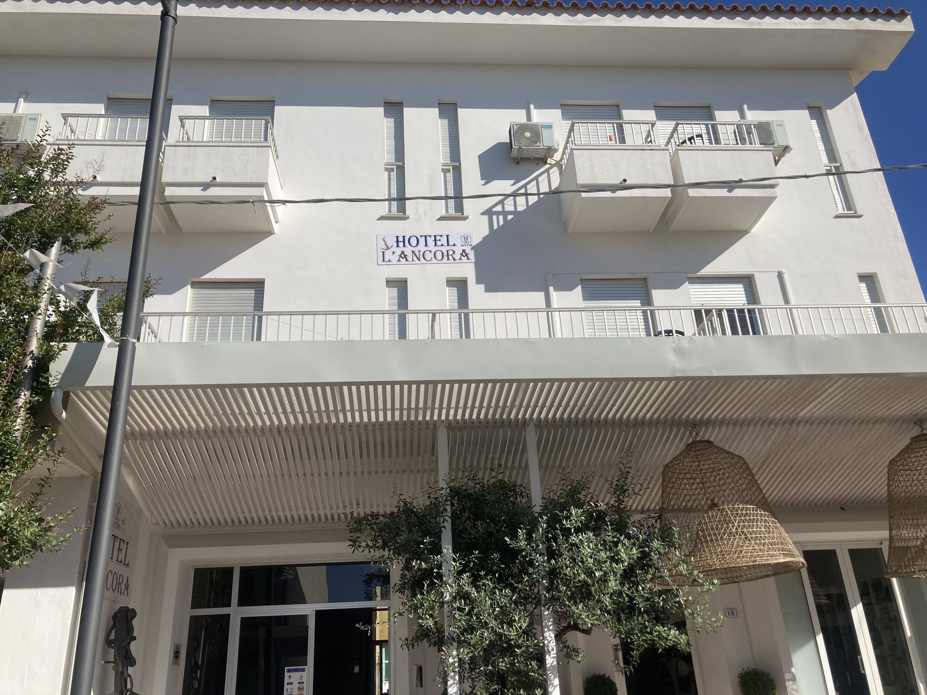 A white building with balconies, a sign reading "HOTEL L'ANCORA," and a covered entrance area with hanging lights.