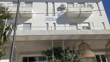 A white building with balconies, a sign reading "HOTEL L'ANCORA," and a covered entrance area with hanging lights.
