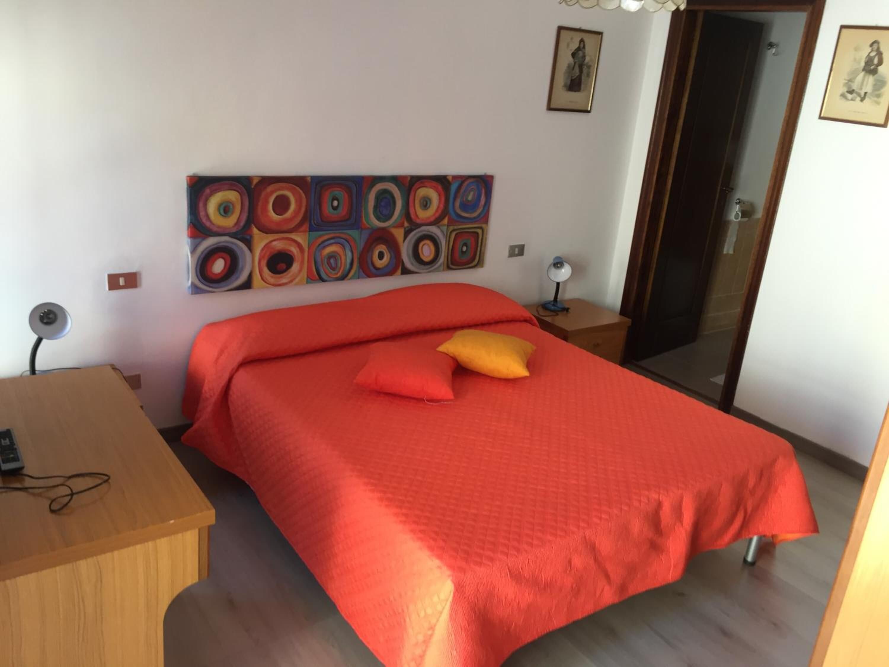 A bedroom with a bed, a desk, a chair, a lamp, and a painting on the wall.