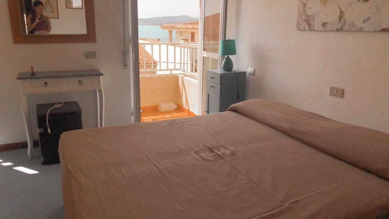 A bedroom with a bed, a dresser, a nightstand, a lamp, and a view of the sea through an open door.
