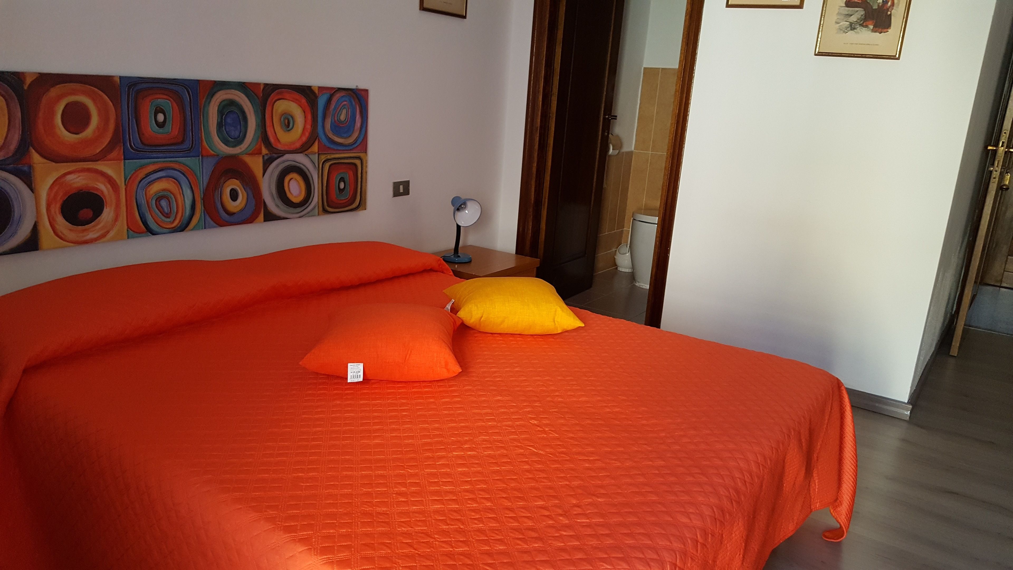 Et værelse med en stor seng, orange sengetøj, et moderne maleri på væggen og et lille natbord med en lampe.