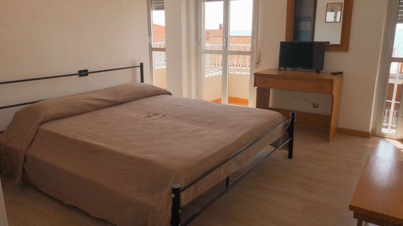 Un dormitorio con cama, escritorio de madera, televisor y puerta corrediza de vidrio que da a un balcón.