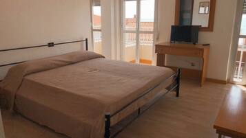 Un dormitorio con cama, escritorio de madera, televisor y puerta corrediza de vidrio que da a un balcón.