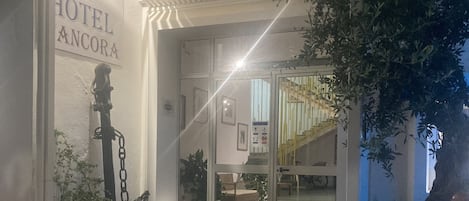 A hotel entrance with a sign reading "HOTEL ANCORA," outdoor seating with chairs, and a staircase leading to the entrance.