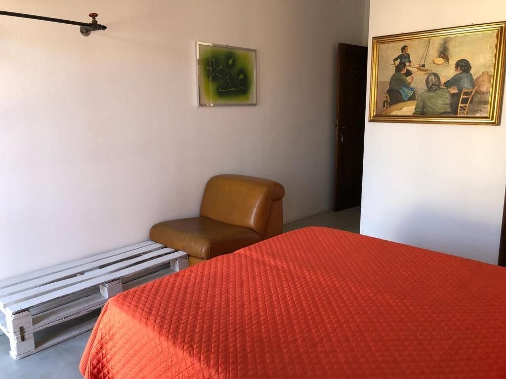Una habitación con una cama, una silla, un banco de madera, un cuadro y una lámpara de pared.