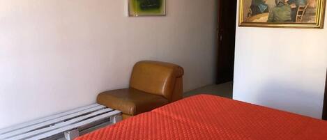 Una habitación con una cama, una silla, un banco de madera, un cuadro y una lámpara de pared.