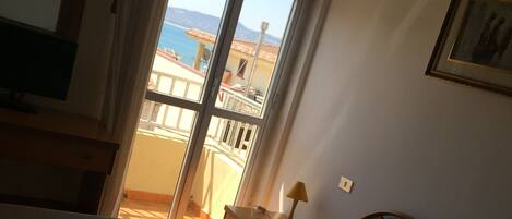 A hotel room with a bed, a desk, a chair, a lamp, a window with a view of the sea, and a framed picture on the wall.