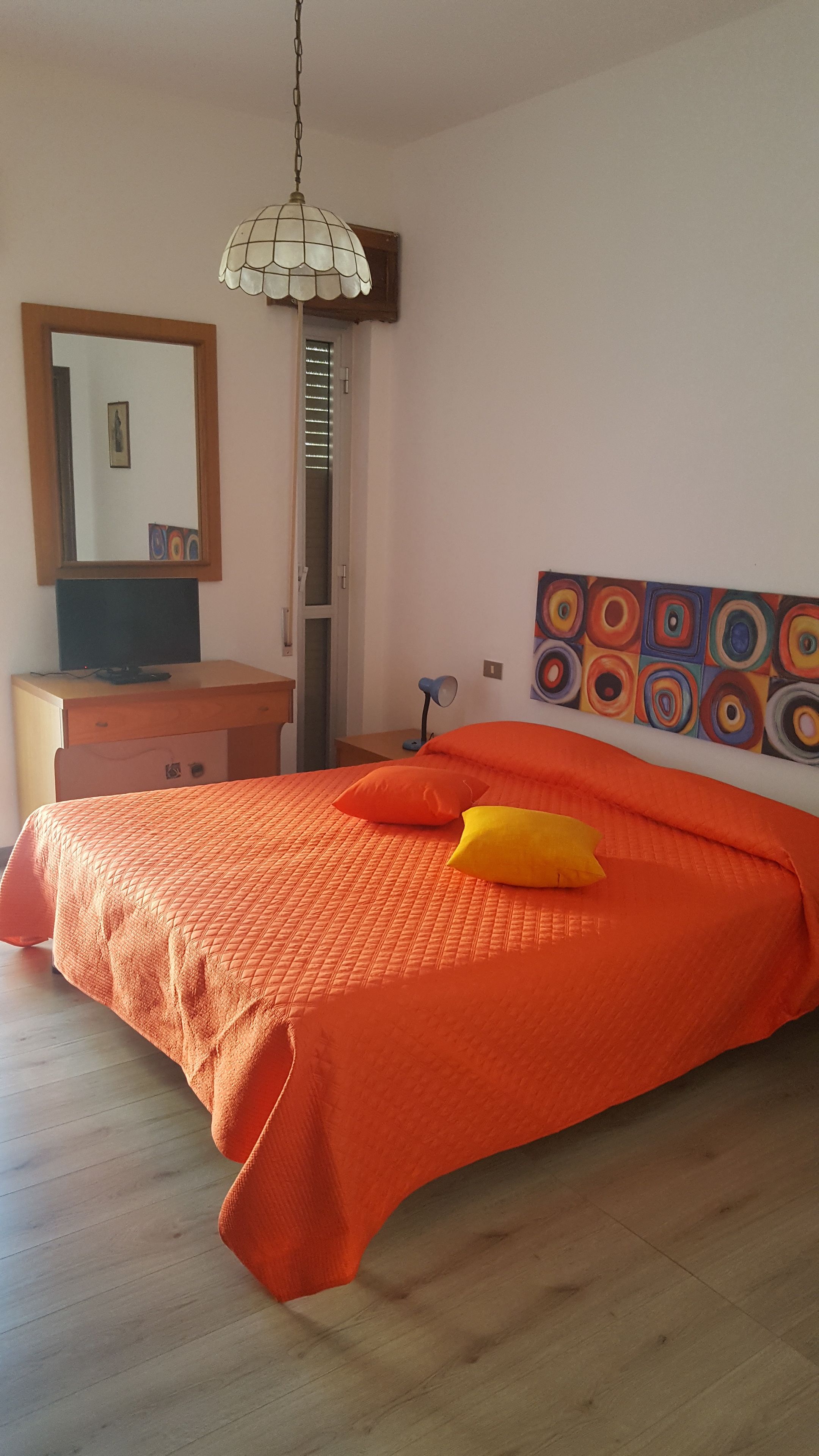 A bedroom with a bed, a desk, a chair, a lamp, a mirror, and a painting on the wall.