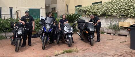Une cour où sont garées quatre motos, alignées les unes à côté des autres, un pot de fleurs et un mur orné de fleurs blanches.