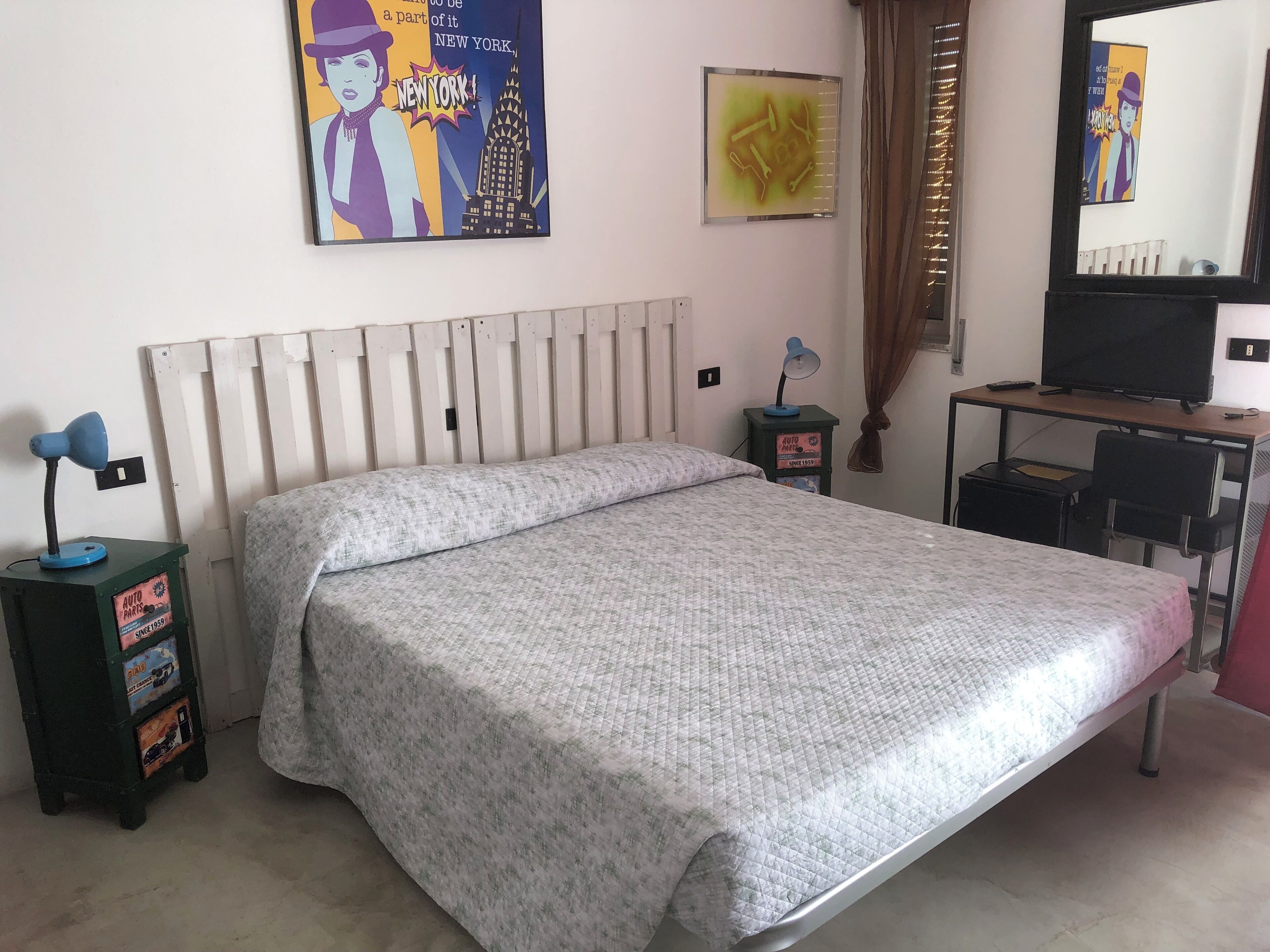 A bedroom with a bed, a desk with a computer, a chair, a lamp, and a poster on the wall.