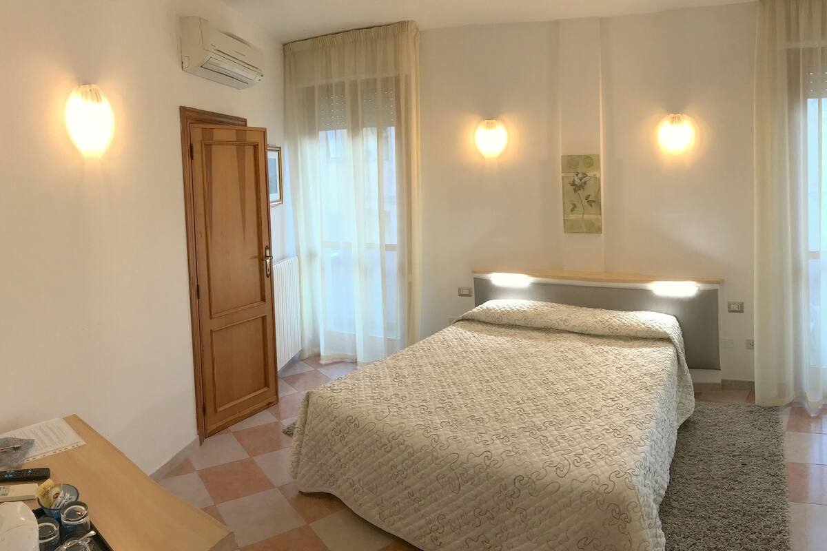 Une chambre à coucher avec un lit, un bureau et une chaise. On y trouve une porte, un radiateur et une fenêtre avec des rideaux.