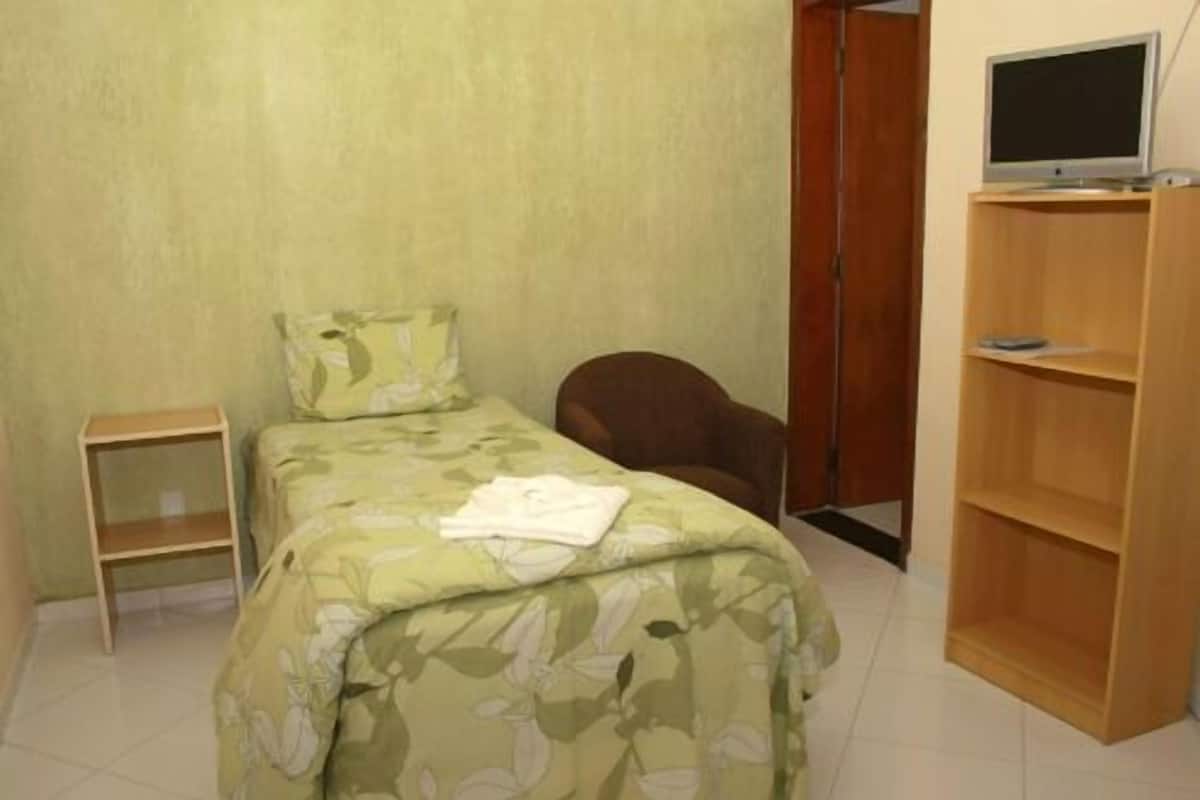 A small bedroom with a single bed, a chair, a TV on a stand, and a wooden shelf unit.