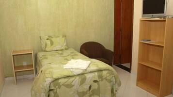 Una camera da letto piccola con un letto singolo, una sedia, un televisore su un supporto e una scaffalatura in legno.
