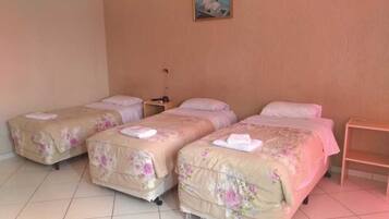 A hotel room with three single beds, each with floral bedding and towels. There is a small bedside table with a lamp and a nightstand with a lamp.