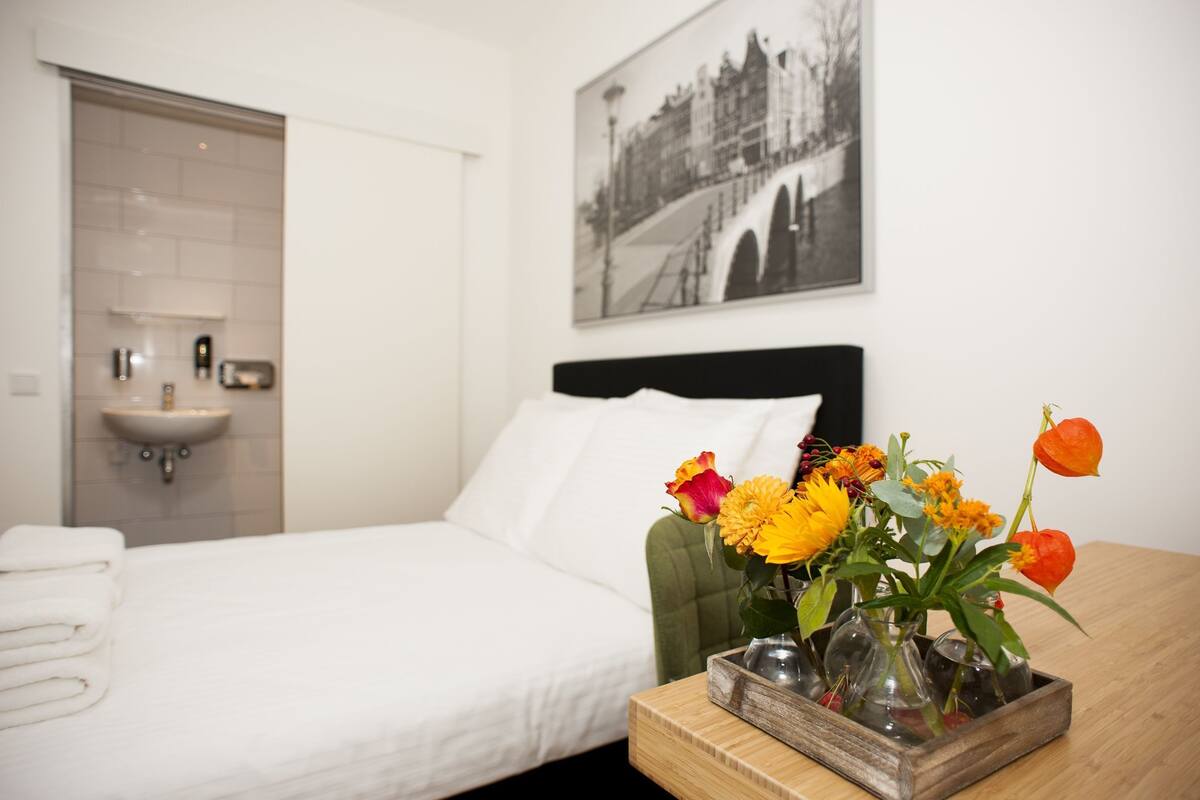 A hotel room with a bed, a bedside table with a vase of flowers, a wall-mounted sink, and a framed picture on the wall.