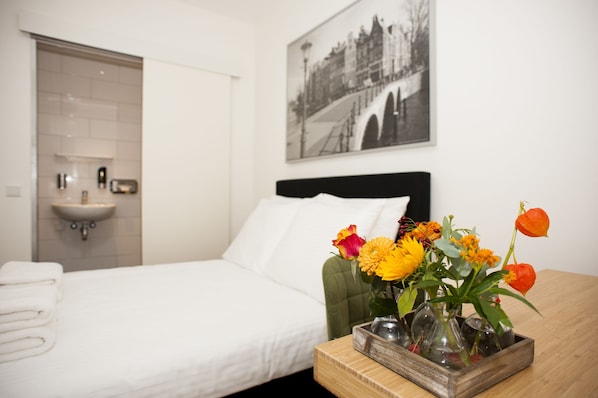 Una habitación de hotel con una cama, una mesita de noche con un florero y una lámpara de pared, un lavamanos empotrado y un cuadro en la pared.