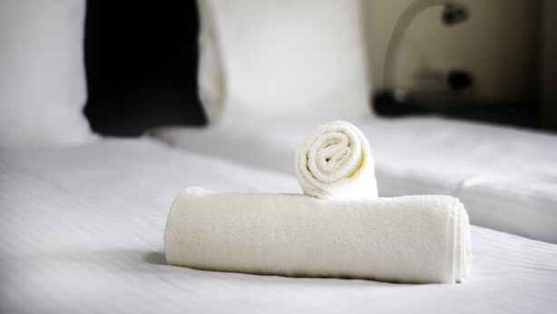 A neatly made white towel on a bed with white bedding and a black headboard. - I Hotel (Amsterdam)