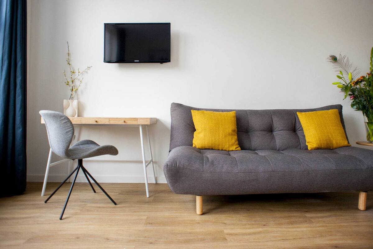 A modern living room with a grey sofa, two yellow cushions, a wooden desk, a grey chair, a black TV mounted on the wall, and a potted plant.