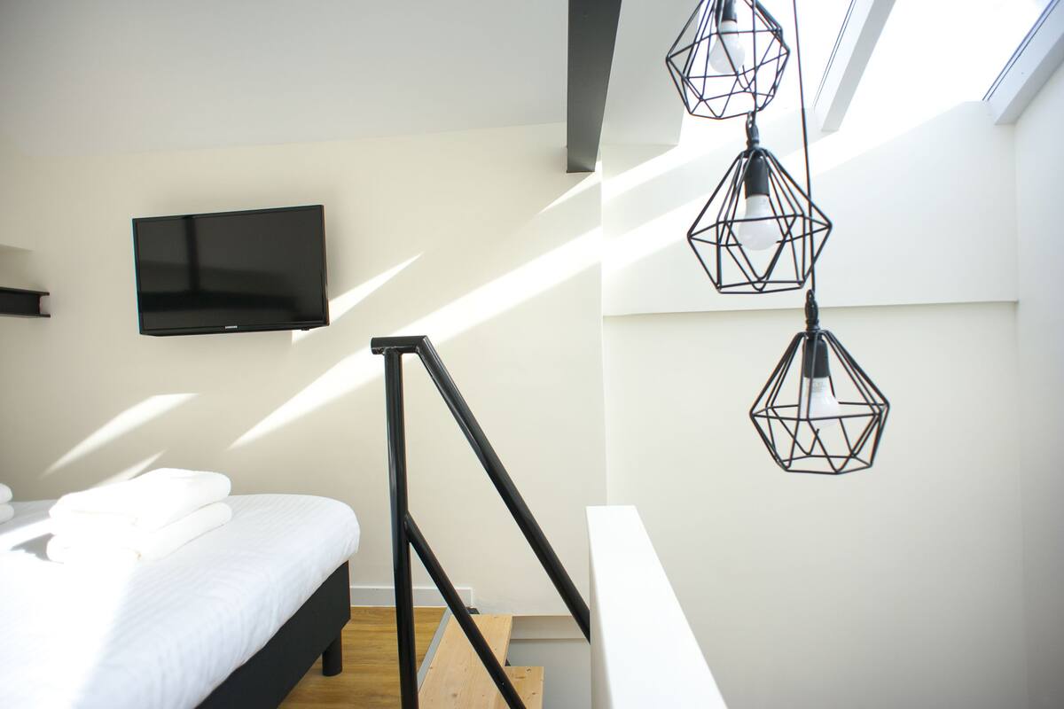 A modern bedroom with a bed, a flat-screen TV, and geometric pendant lights.