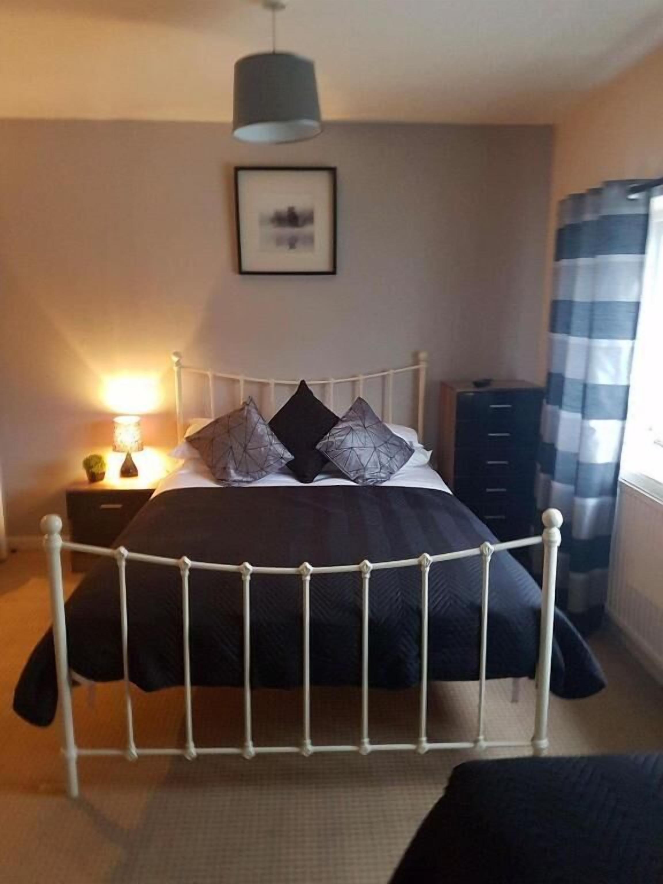 A bedroom with a bed, a nightstand, a lamp, a dresser, and a window with curtains.
