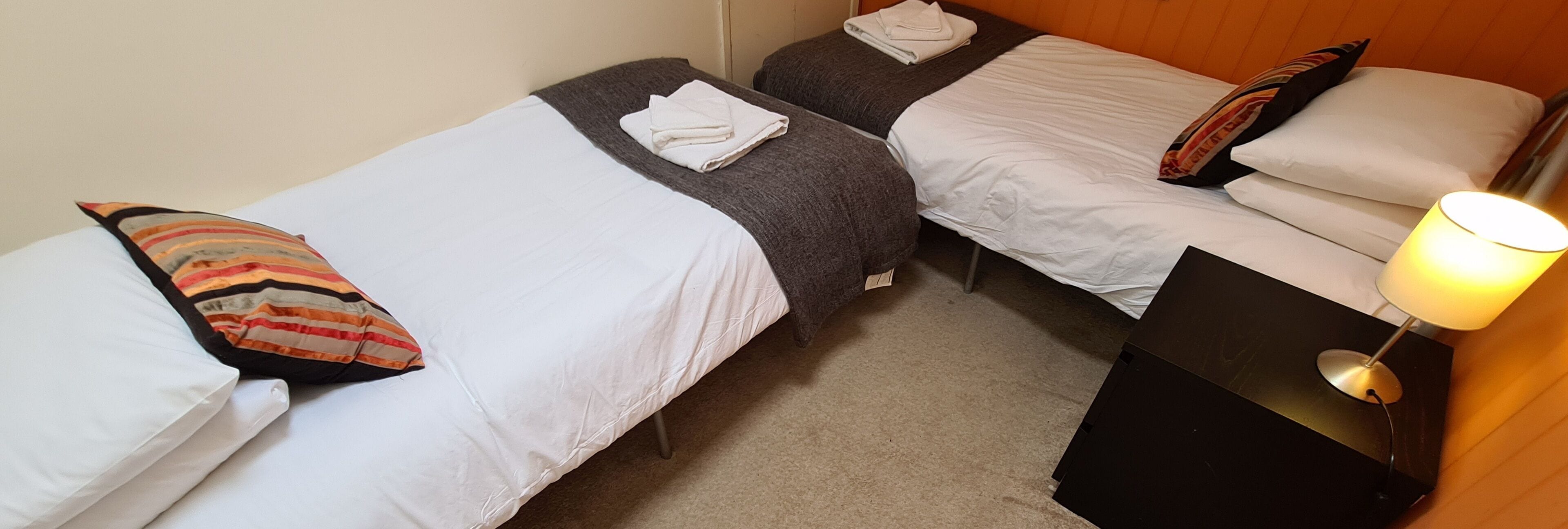 A hotel room with two single beds, each with a pillow and a folded towel. There is a nightstand with a lamp on the right side.