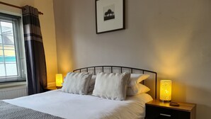 A neatly made bed with white linens and striped pillows, a bedside table with a lamp, and a window with curtains.