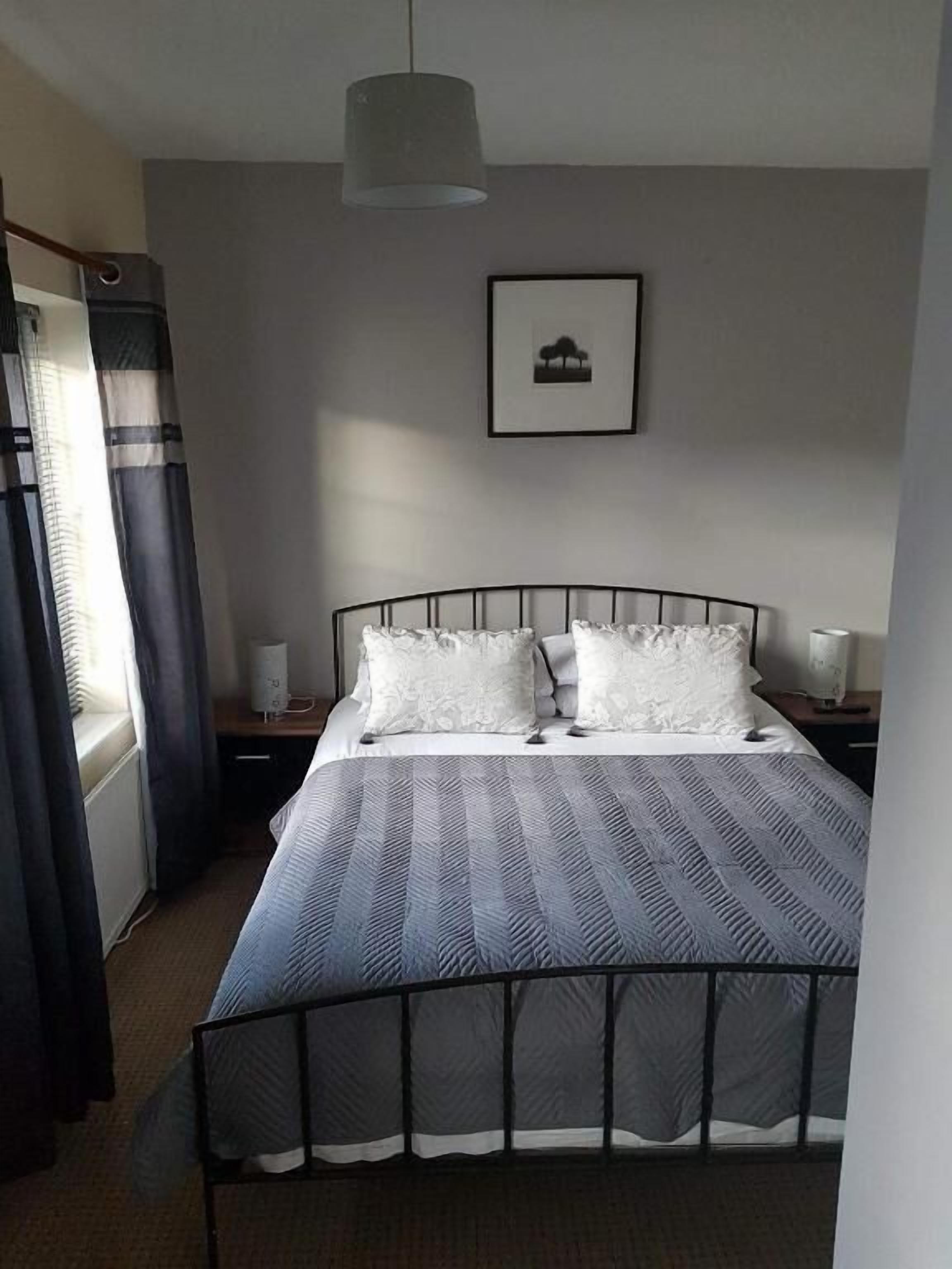 A bedroom with a bed, a bedside table, a lamp, a picture on the wall, and a window with curtains.