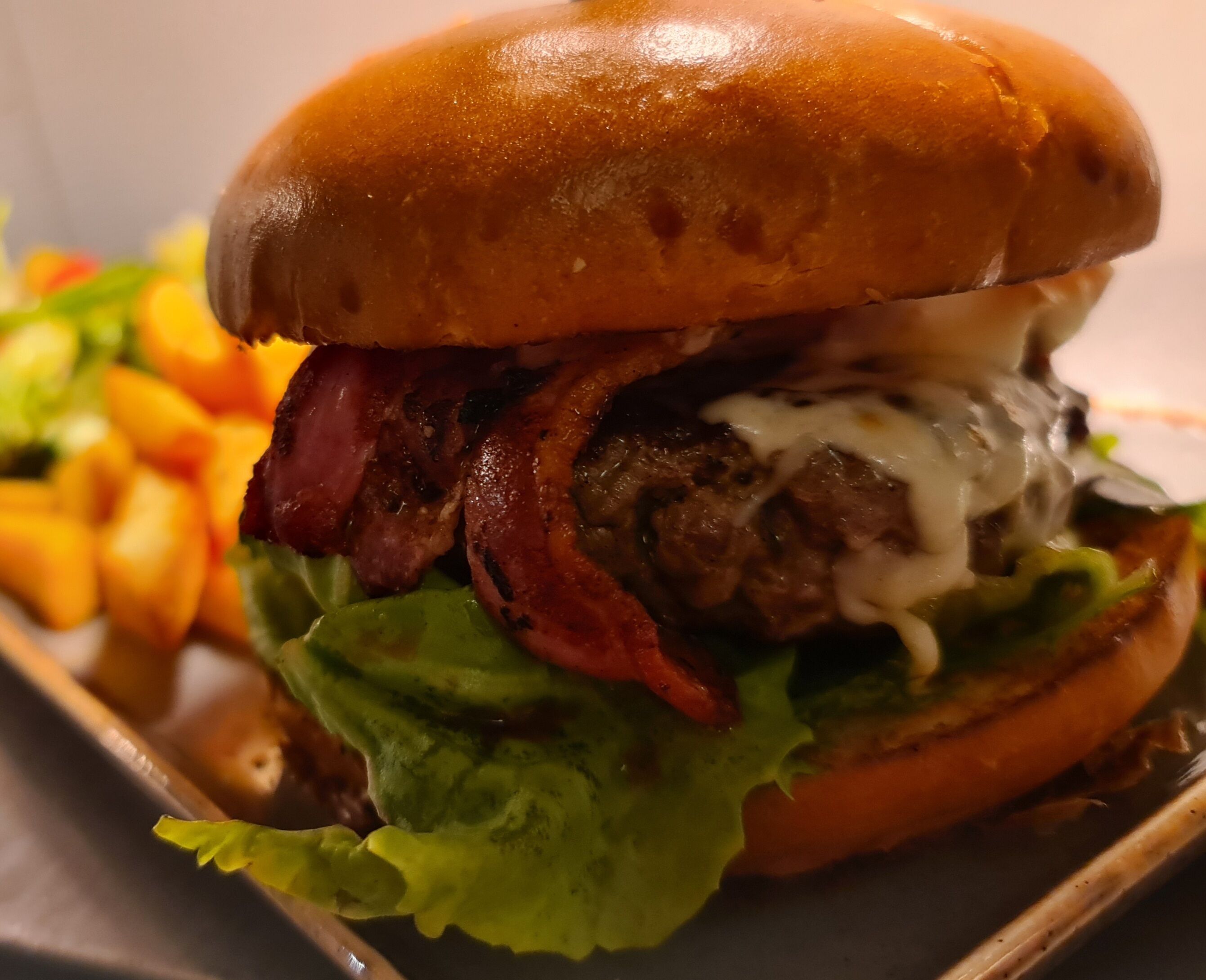 A close-up of a burger with a sesame seed bun, bacon, lettuce, and a creamy sauce.