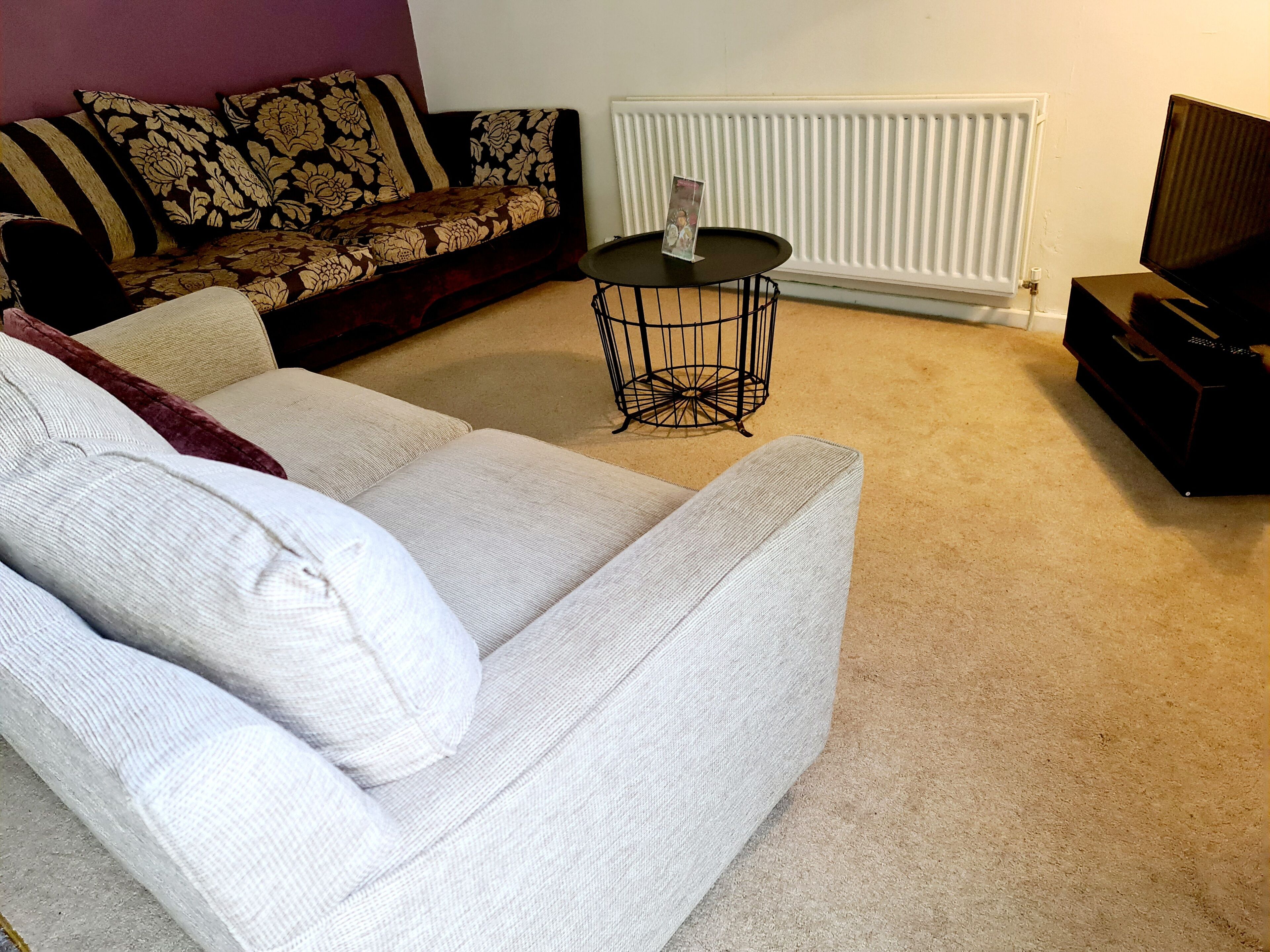 A living room with a white sofa, a patterned sofa, a small black table with a magazine, a radiator, and a TV stand.