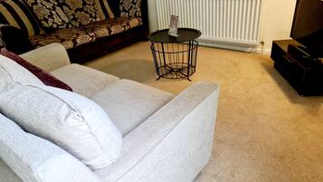 A living room with a white sofa, a patterned sofa, a small black table with a magazine, a radiator, and a TV stand.