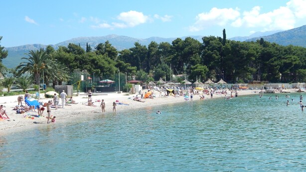 A beach with people swimming and sunbathing, surrounded by trees and mountains in the background. - Apartments Milenka (Split)