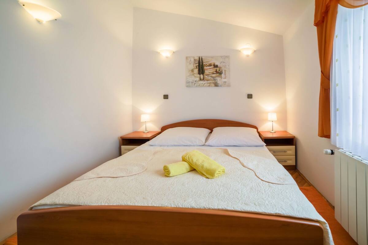 Ein ordentlich bezogenes Bett mit weißer Daunendecke und zwei gerollten Handtüchern, zwei Nachttischlampen, ein kleiner Tisch mit Lampe und ein Bild an der Wand.