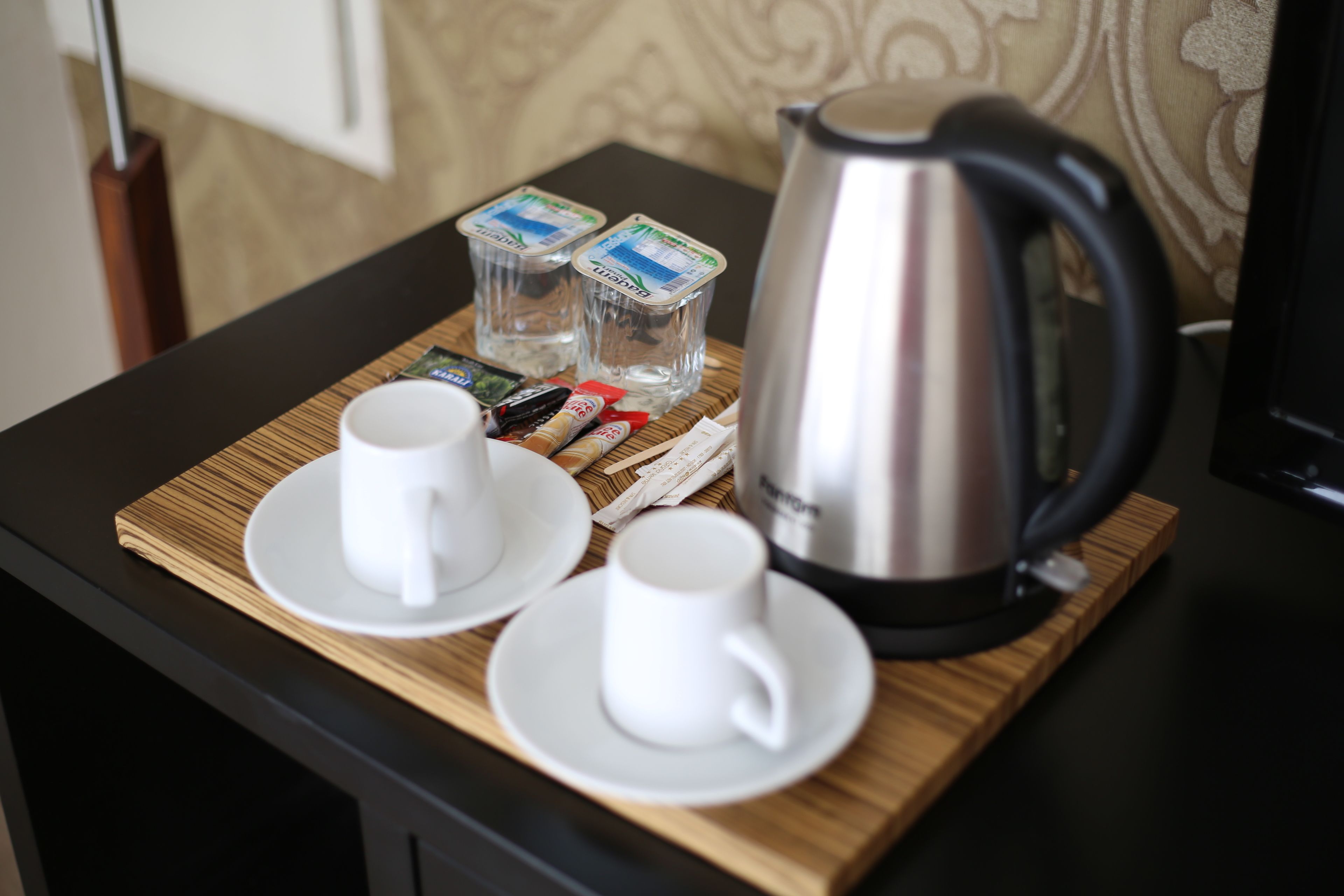 A breakfast tray with two white cups, a silver electric kettle, and two containers of yogurt on a wooden table.